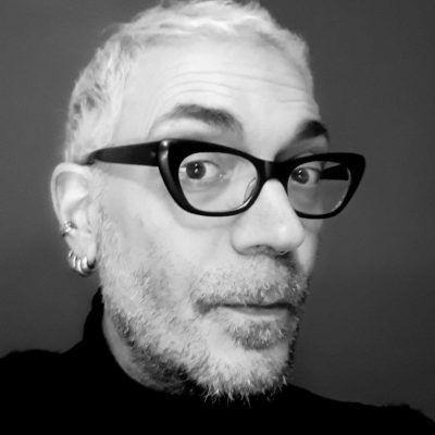 Foto in bianco e nero con il volto dell'autore uomo con capelli bianchi, corti, barba e occhiali neri. Indossa una maglia nera