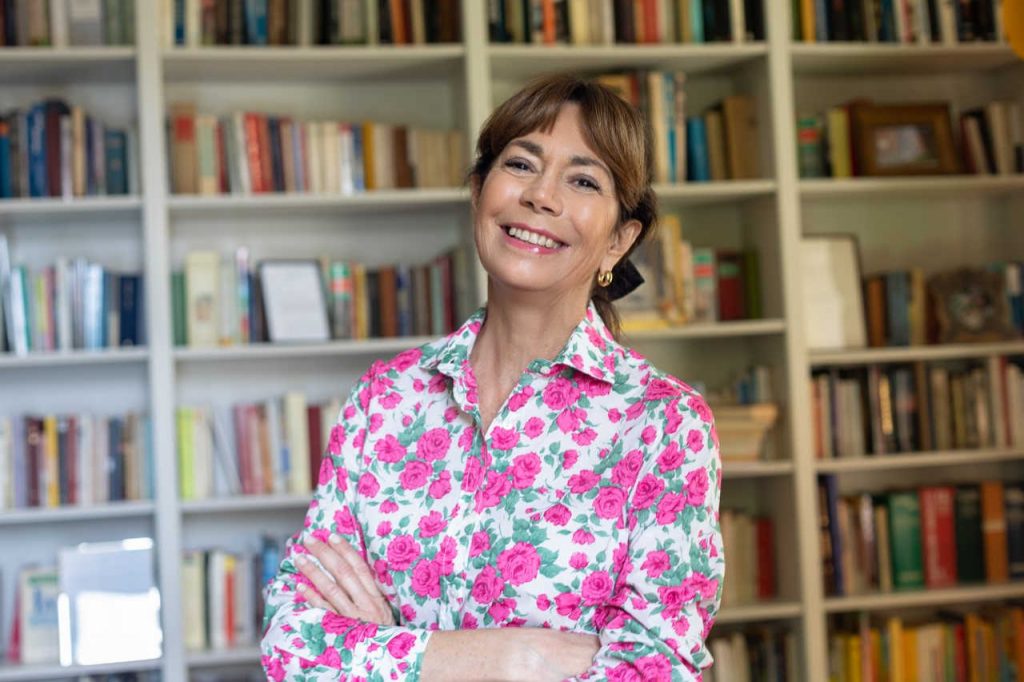 Donna sorridente, capelli castani corti e camicetta bianca con fiori rosa. Si trova in una stanza con dietro una libreria