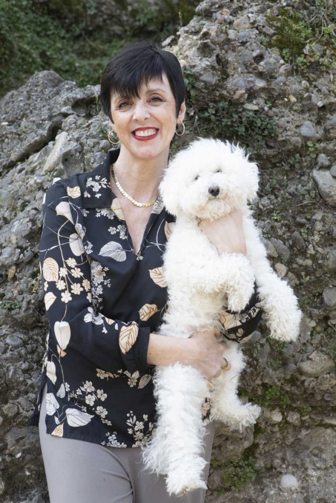 Donna sorridente capelli scuri corti con in braccio un cane barboncino bianco. Indossa una camicetta blu con delle stampe e pantaloni grigi