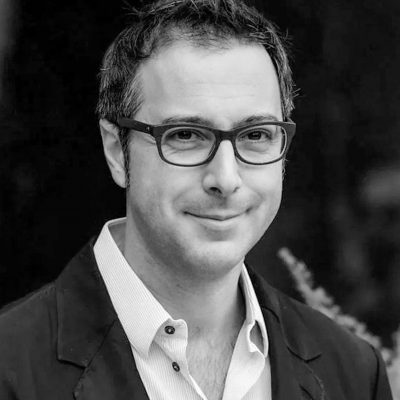 Foto di uomo sorridente in bianco e nero. Capelli scuri con occhiali. Giacca scura e camicia bianca
