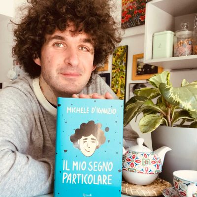 Autore Libro - Ragazzo con capelli mori ricci , maglia grigia che mostra il suo libro