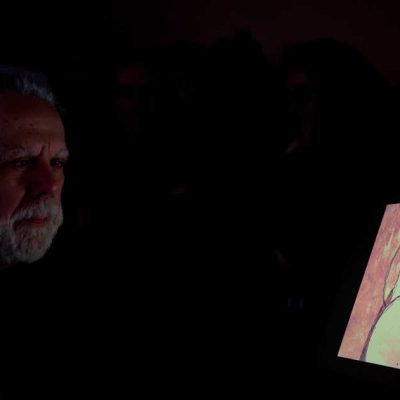 Foto con sfondo nero con l'autore uomo capelli e barba bianca che guarda l'immagine del fumetto Bobo: faccia di uomo con grandi occhiali e grande naso