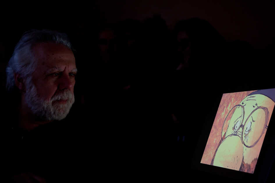 Foto con sfondo nero con l'autore uomo capelli e barba bianca che guarda l'immagine del fumetto Bobo: faccia di uomo con grandi occhiali e grande naso