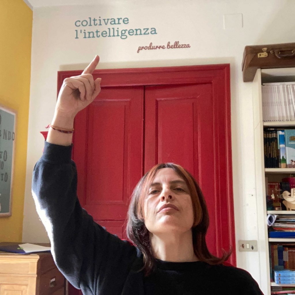 Foto di donna in primo piano, entro una stanza. Ha un braccio alzato e un dito che indica una scritta nel muro "coltivare l'intelligenza", sotto c'è una porta rossa. La donna è castana con divisa nel mezzo e indossa una maglia blu.