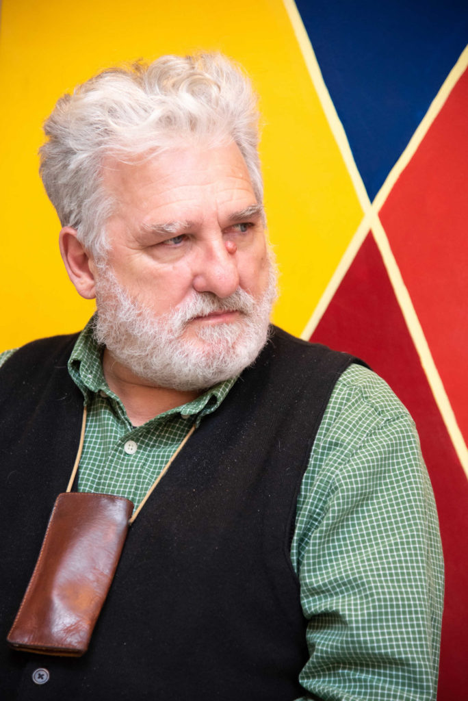 Uomo con barba e capelli bianchi che guarda a sinistra. Indossa camicia verde, gilet nero e al collo ha un portaocchiali col marrone