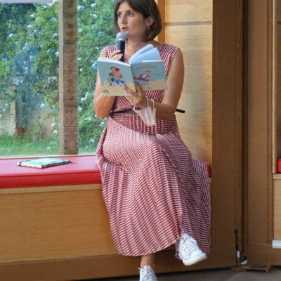 Donna con microfono in mano che legge un libro. Ha capelli a caschetto castani e indossa un vestino rosa