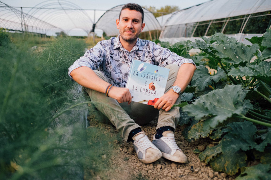 Uomo seduto in una serra con un libro in mano in mezzo a piante di finocchi e zucchine