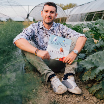 Uomo seduto in una serra con un libro in mano in mezzo a piante di finocchi e zucchine