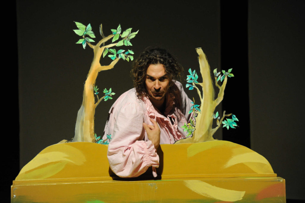 Uomo che recita a teatro, poggiato su una sagoma gialla con due piccoli alberi ai lati.